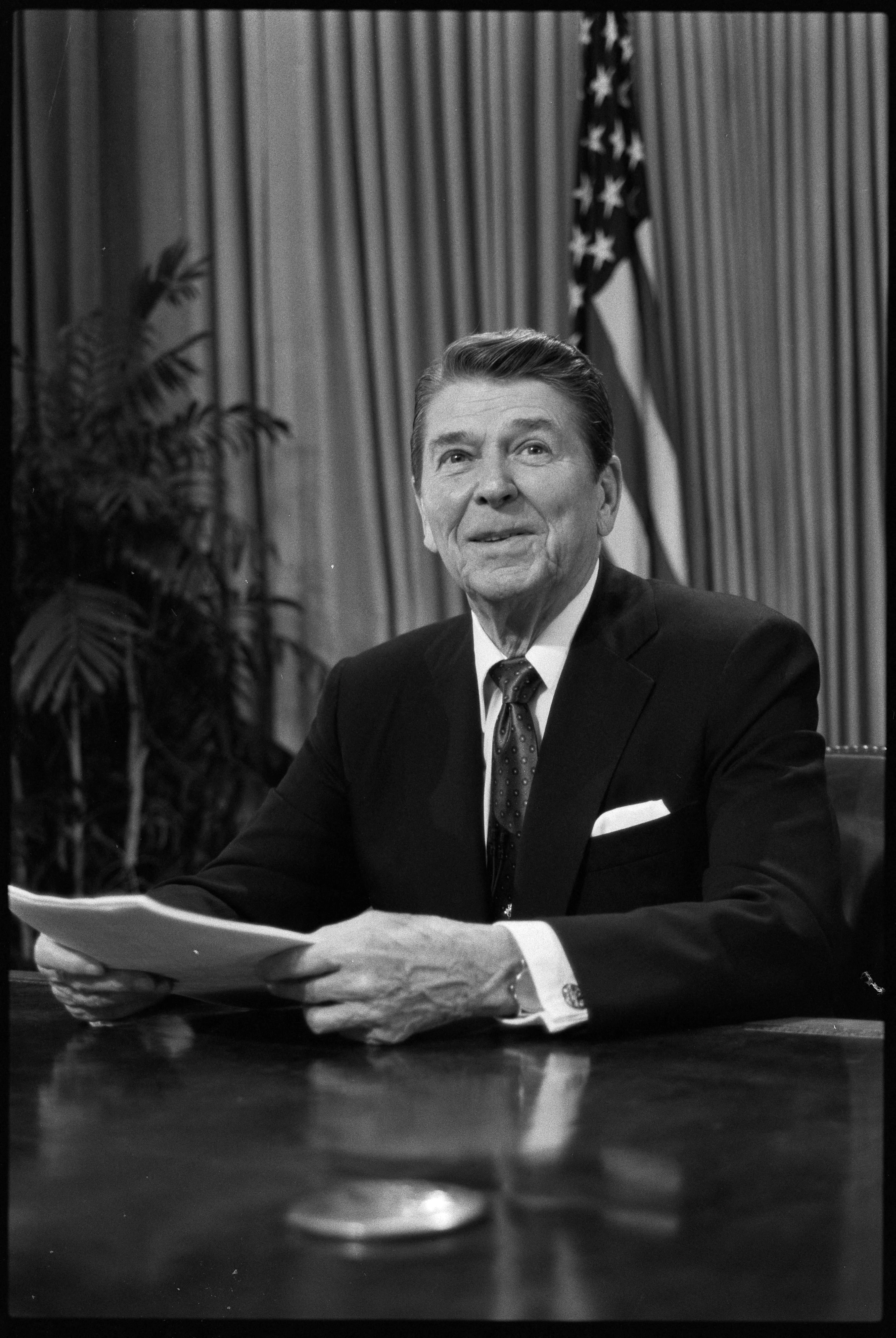 A black and white photo of Reagan sitting in the Oval Office wearing a suit and tie.