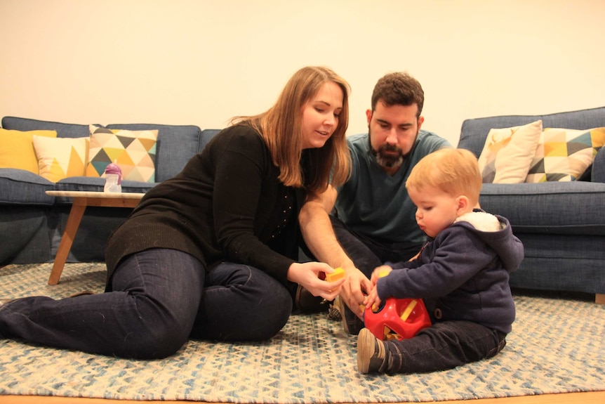 A man and woman sit on a rug in a living room and play with a toddler.