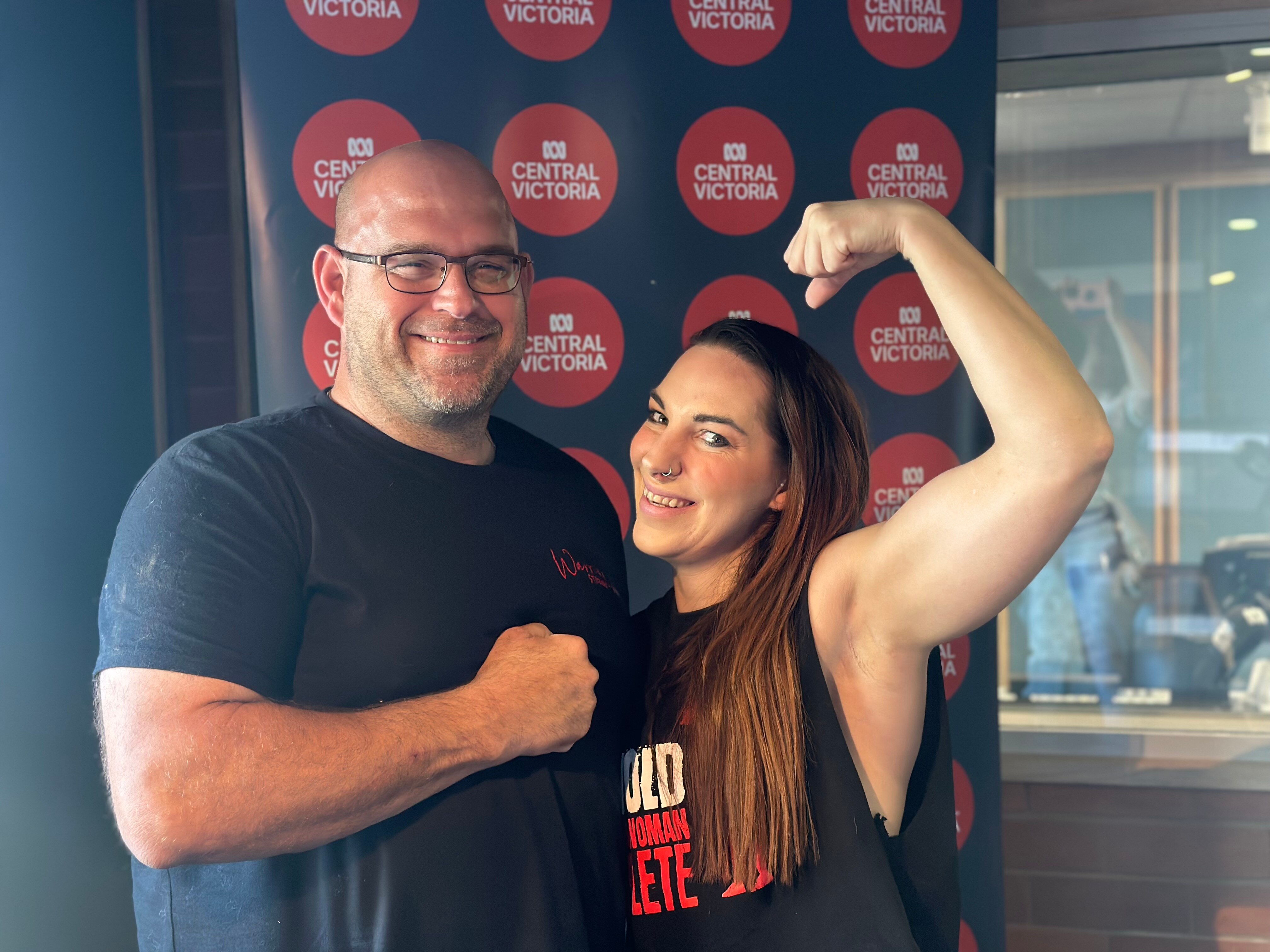 A man and a woman in gym gear smile at the camera. The woman poses her biceps.