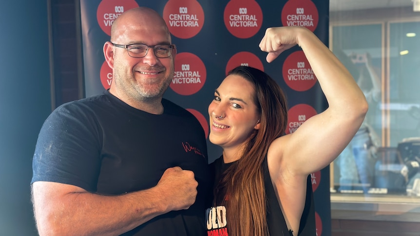 A man and a woman in gym gear smile at the camera. The woman poses her biceps.