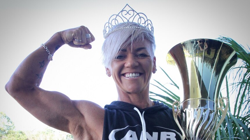 Woman stands smiling with muscles flexed and holding trophy.