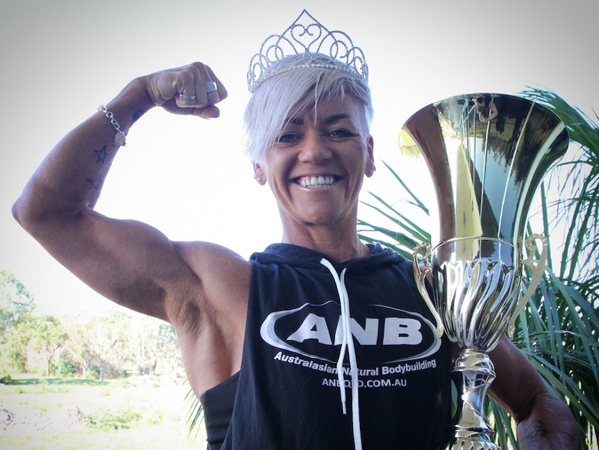 Woman stands smiling with muscles flexed and holding trophy.