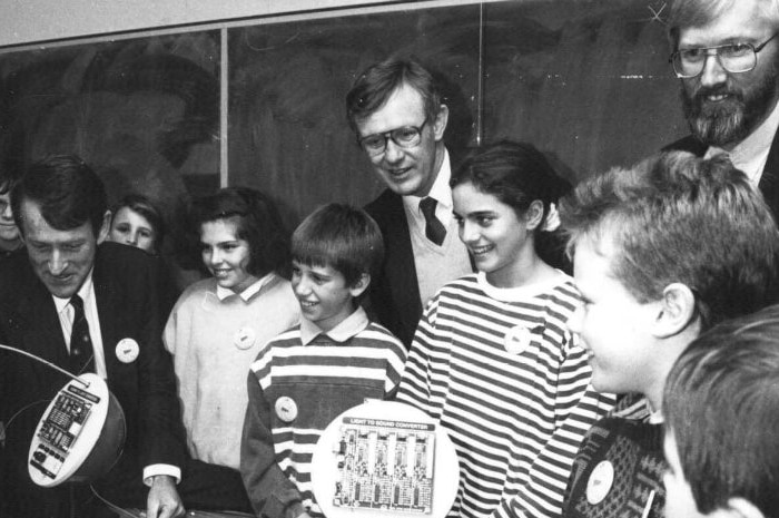 Three adult men stand behind curious children looking at strange scientific instruments, it's a black and white photo