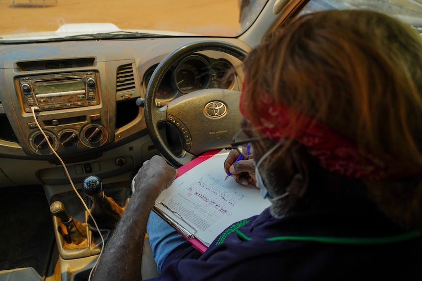View from over the shoulder of a man writing on a clipboard in the driver's seat of a car.