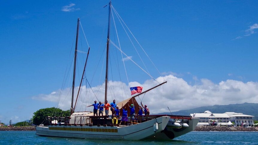 Nine crew member stand celebrating on a tradition boat in Samoa harbour flying the Samoa flag. 