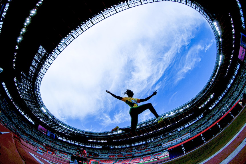 A Jamaican athlete flying through the air during the long jump final in Tokyo stadium, shot from below so the sky is above them