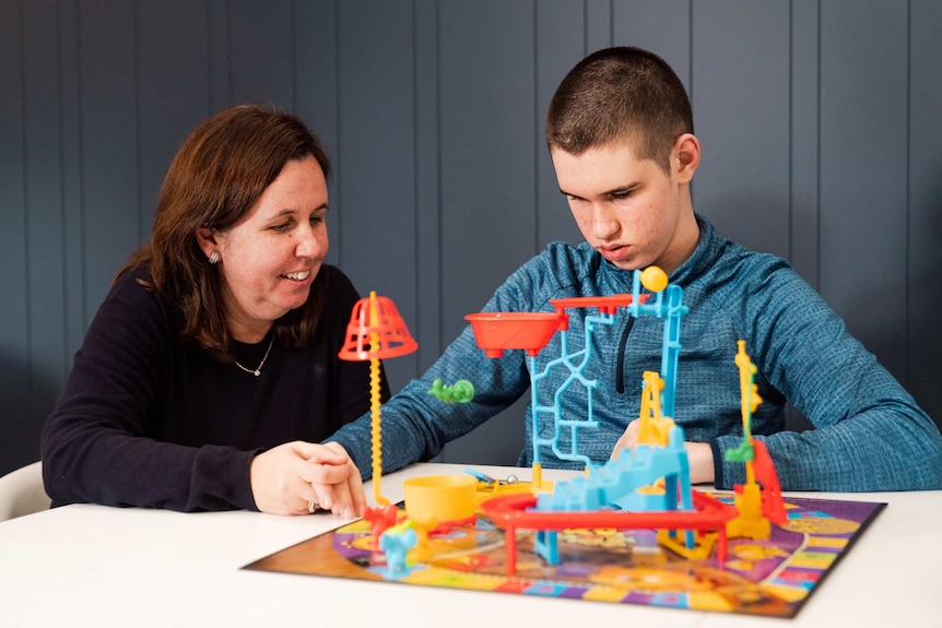 A teenage boy sits at a table playing a children's board gane as his mother sits alongside him smiling.