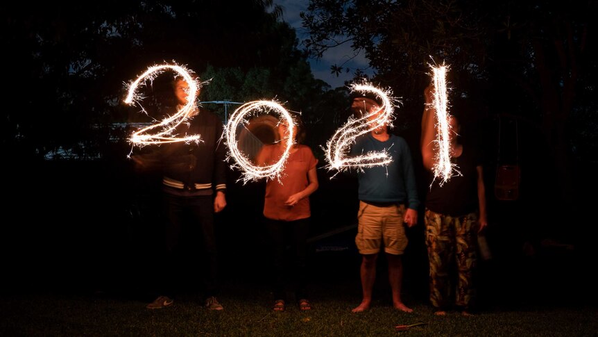 Four people each holding a sparkler write '2021' with the sparklers at night