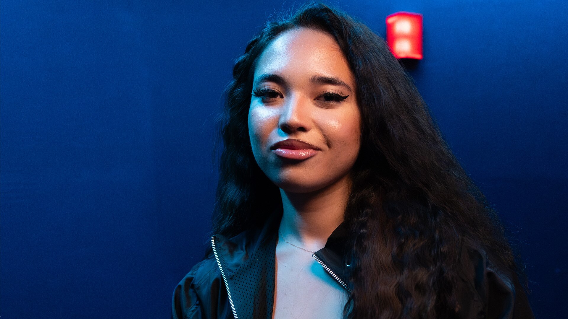 A woman in long dark hair in a black jacket and white top smiles in a blue room