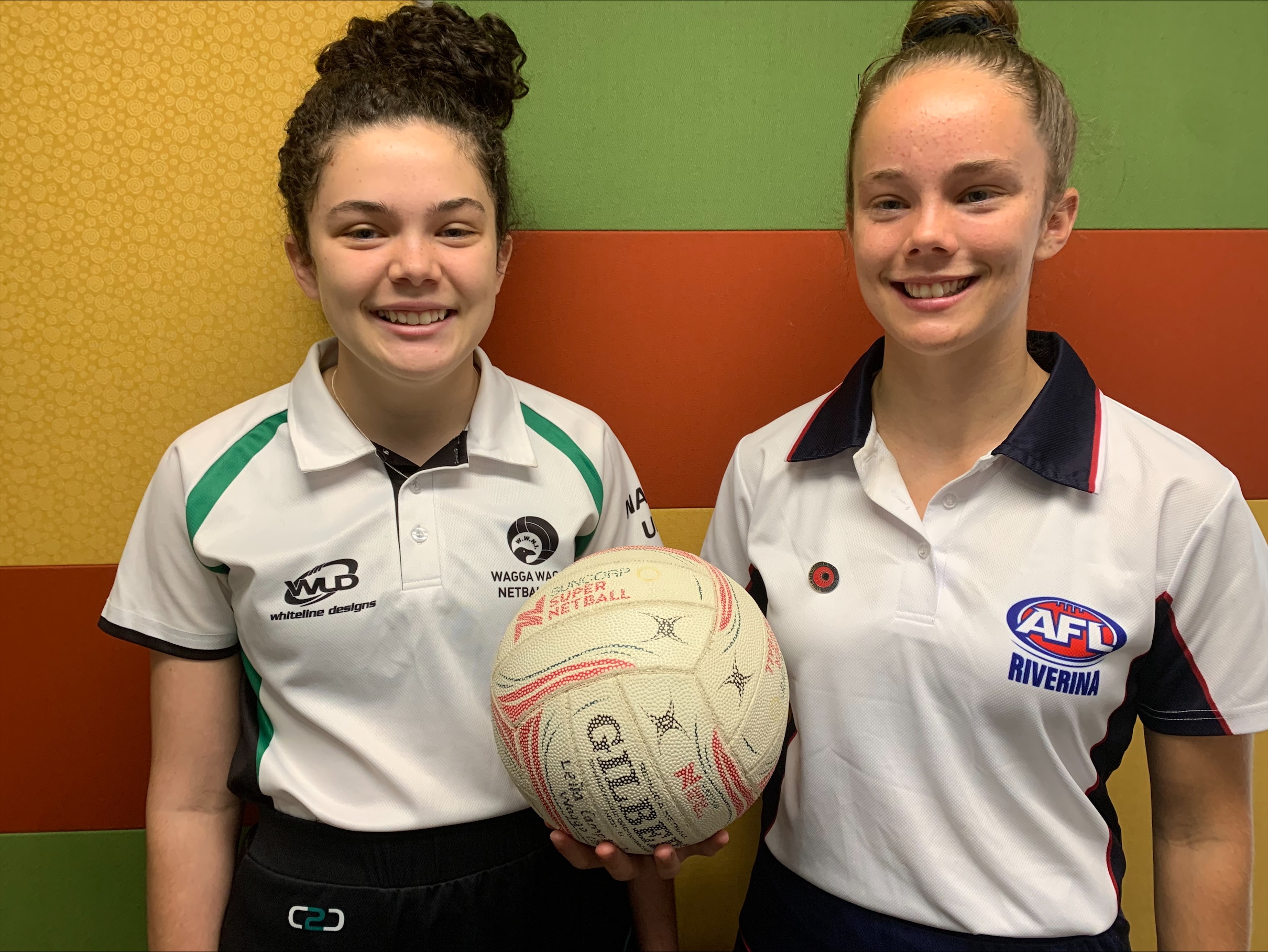 Two young woman standing side by side, holding a netball in one hand each.