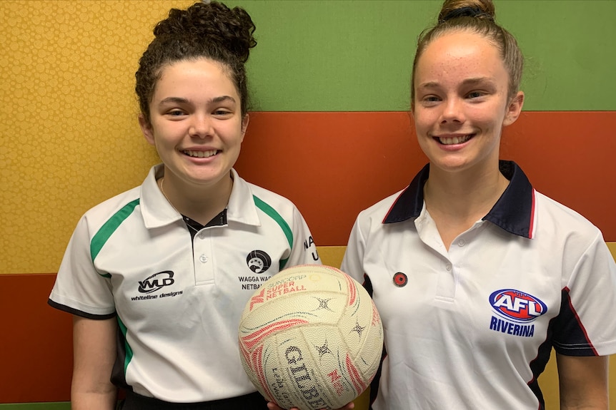 Two young woman standing side by side, holding a netball in one hand each.