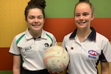 Two young woman standing side by side, holding a netball in one hand each.