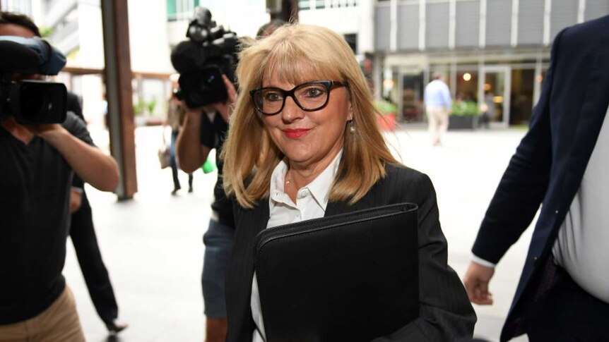 A woman with straight blonde hair, black glasses, a black jacket and a white shirt, is filmed by TV cameras.