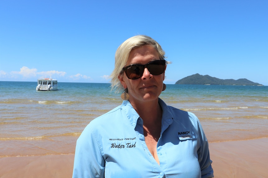 A woman in a blue shirt stands on a beach with a boat and Dunk Island in the background