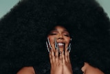 A woman with very long black fingernails holds her hands to her face