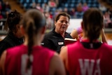 A woman in a dark shirt speaking to Netbal players in red shirts.