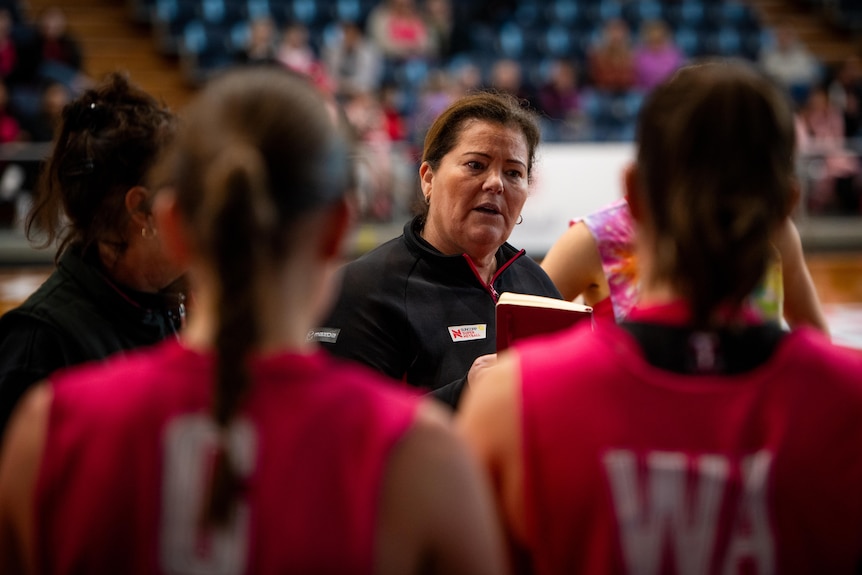 A woman in a dark shirt speaking to Netbal players in red shirts.