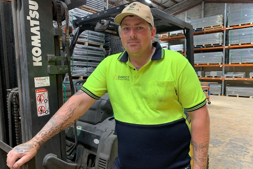 A man in a yellow high-vis shirt and a cap stands beside a forklift in a warehouse.
