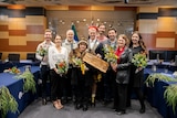A group of people hold native flowers and a piece of wood with writing on it.  