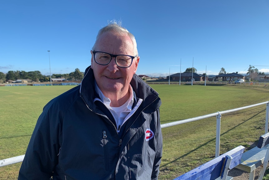 a man wearing glasses and a winter jacket standing beside an australian rules football ground