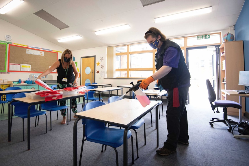 Teaches disinfect primary school desks in a classroom while wearing masks.