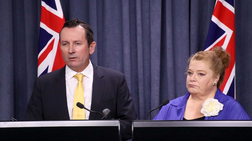 A man and a woman looking sad, standing behind a lectern with Australian flags behind them.