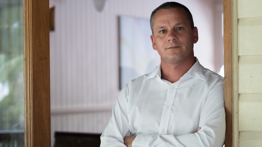 A man in a white shirt crosses his arms as he leans against the lintel of a property doorway.