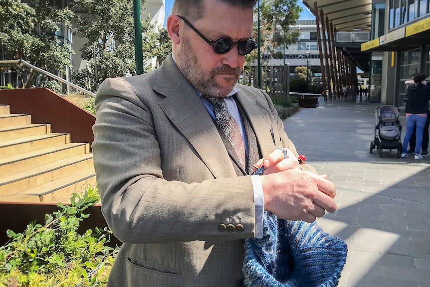 Wolf wears a grey suit and dark, round sunglasses as he looks down with concentration at a brioche garment he is knitting.