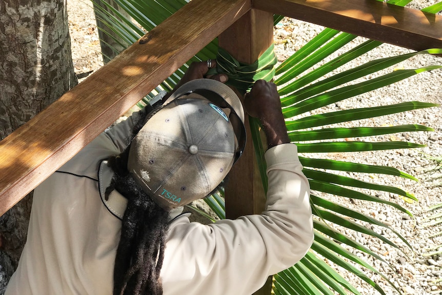 Man with workwear weaving coconut leaves around a pole.