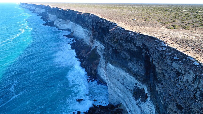 Tall cliffs with ocean waves and tiny person and vehicle on top