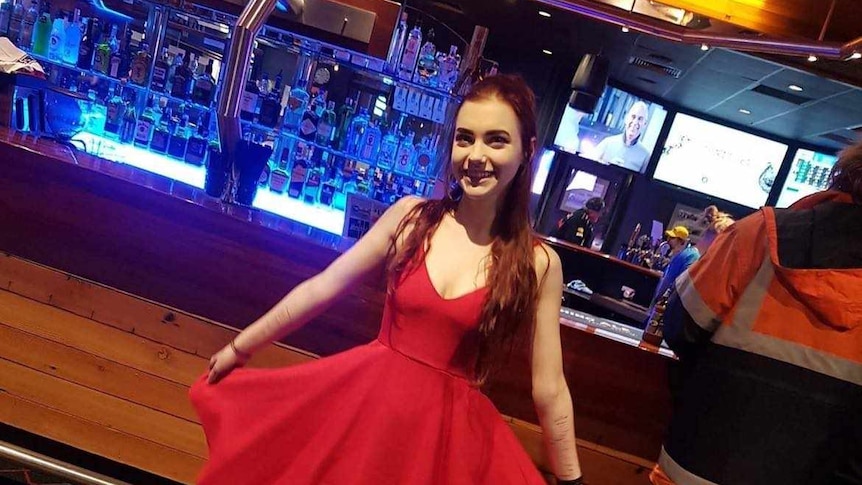 A young woman with brown hair stands in front of a wooden drinks bar. She is wearing a long red dress. 
