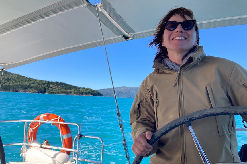 A smiling woman with dark hair, wearing a jacket and sunglasses, helms a small boat on a sunny day.