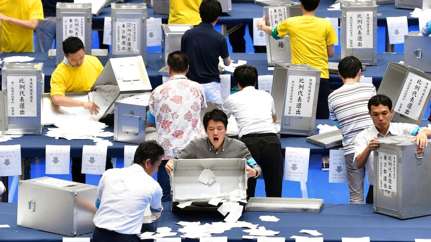 A bird's eye view shows three rows of Japanese election officials opening silver ballot boxes onto blue trestle tables.