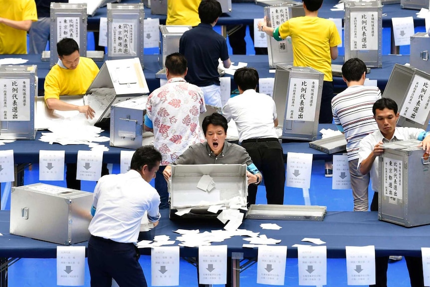A bird's eye view shows three rows of Japanese election officials opening silver ballot boxes onto blue trestle tables.