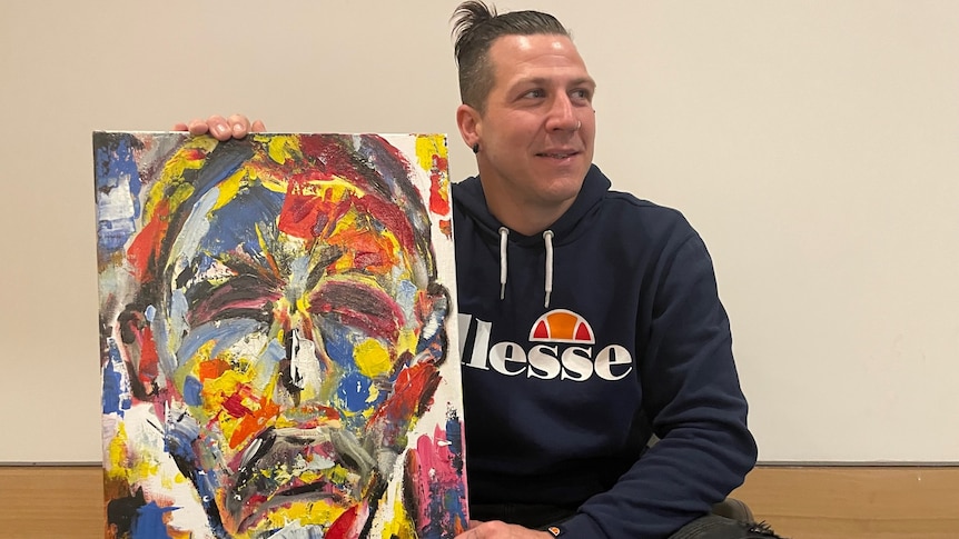 A man in a blue hoodie and his hair tied back in a bun, holds an abstract acrylic self-portrait. 