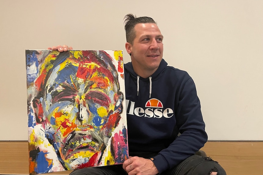 A man in a blue hoodie and his hair tied back in a bun, holds an abstract acrylic self-portrait. 