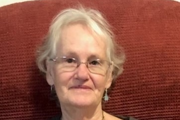 An elderly woman sits on a chair and smiles at the camera