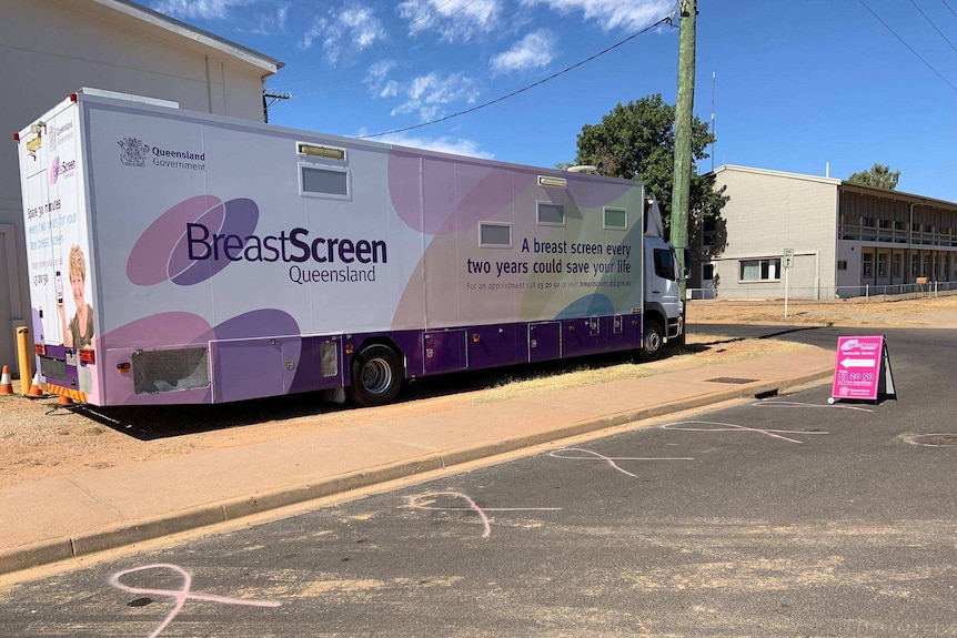 A BreastScreen bus parked on the footpath with a pink sign about how to book, with pink breast cancer ribbons on the roadside