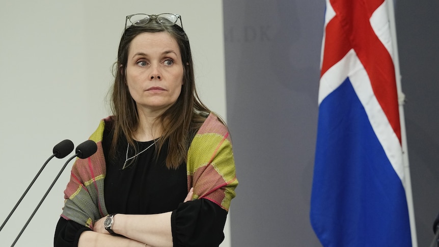 A middle-aged woman crosses her arms and looks unamused in front of the Icelandic flag.