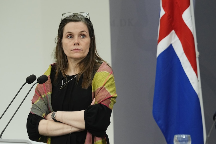 A middle-aged woman crosses her arms and looks unamused in front of the Icelandic flag.