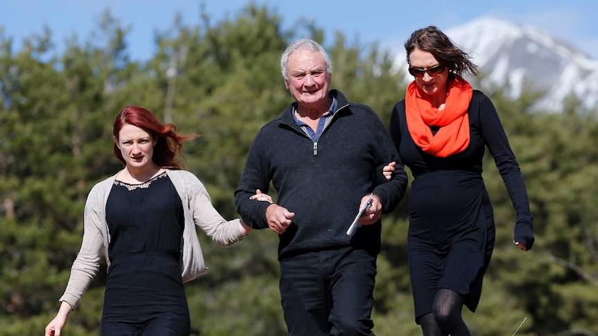 A grieving man is supported by two women with snow-capped mountains in the background