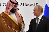 Vladimir Putin and Mohammed bin Salman both have half-hearted smiles on their faces as they shake hands and look at each other.