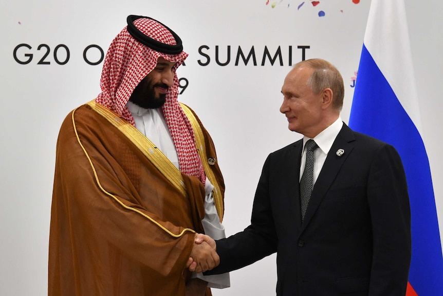 Vladimir Putin and Mohammed bin Salman both have half-hearted smiles on their faces as they shake hands and look at each other.