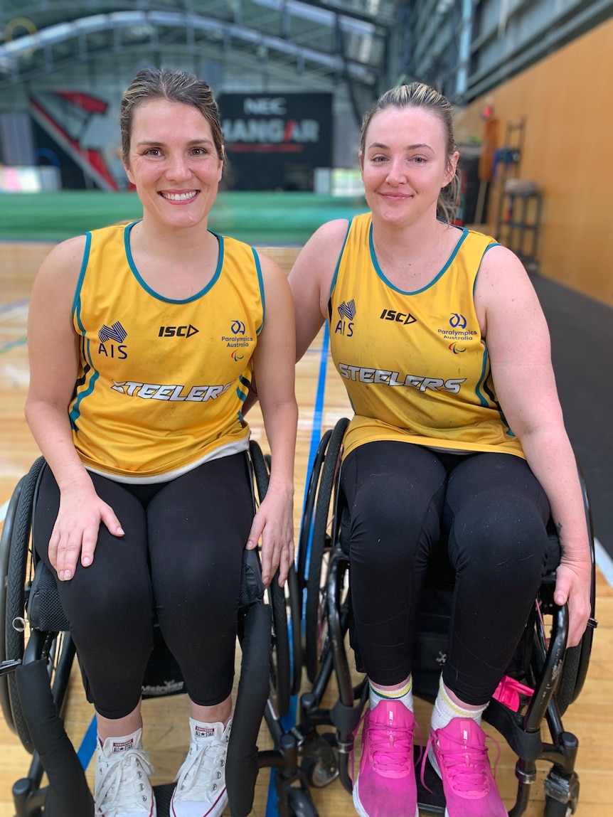 Two Australian female wheelchair rugby players sit in their chairs and smile at the camera.