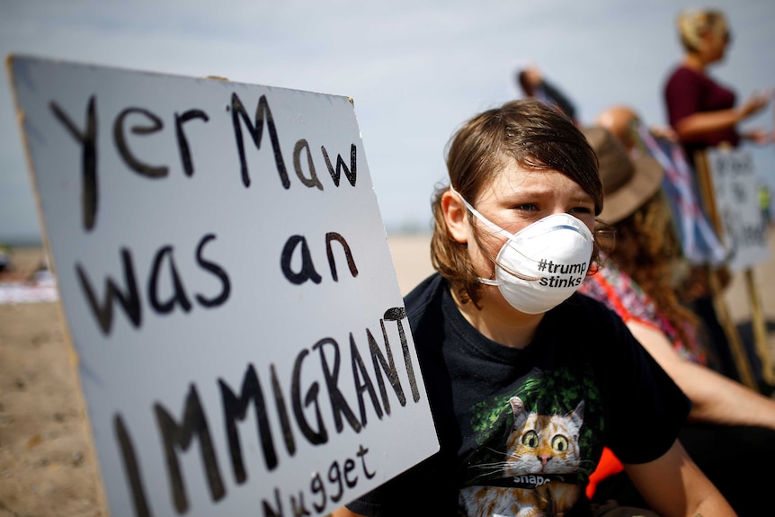 A teen boy sits on a beach holding a sign that says "Yer Maw was an Immigrant nugget", wearing a mask that says "#Trumpstinks"