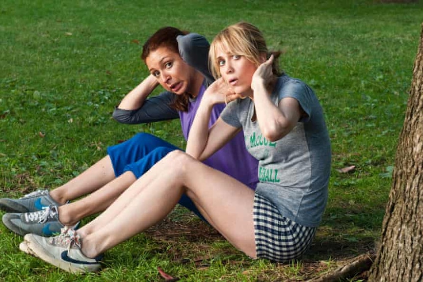 Two women sit on grass doing stomach crunches and peering behind a tree