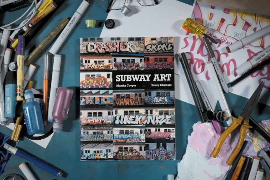 A photobook featuring graffiti sits on desk surrounded by texters and other drawing utencils.
