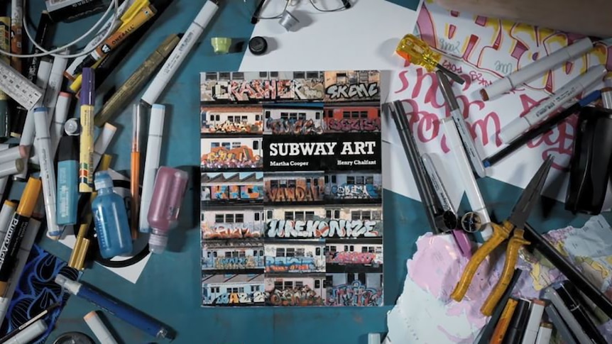 A photobook featuring graffiti sits on desk surrounded by texters and other drawing utencils.