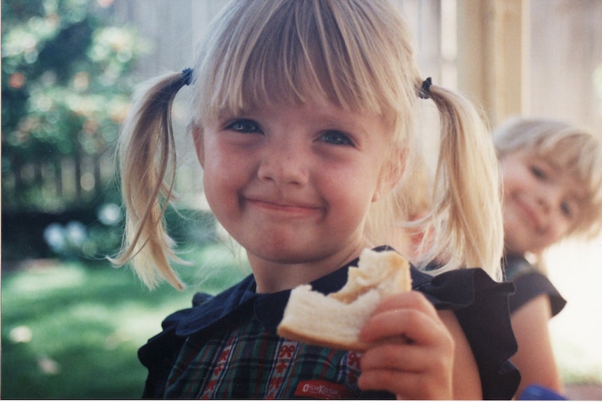 Young girl with blonde brown hair smirking, holding a bitten off sandwich, young kid in background.