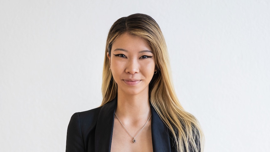 Emily, with blonde hair over her shoulders, stands in front of a white background wearing a black blazer and white shirt